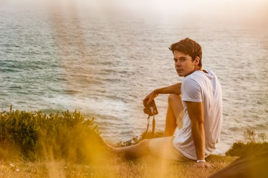 A young man in a white t-shirt sits on a grassy cliff, enjoying the ocean view during sunset.