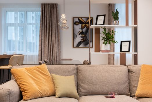 Light room interior with soft couch with pillows and glass of spilled wine near shelves and counter with chairs near window with curtains