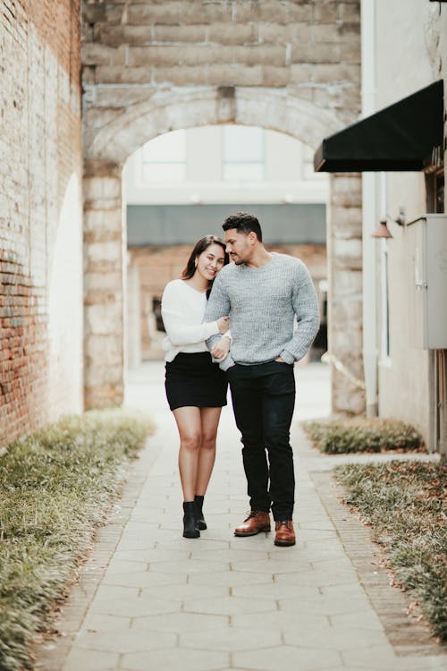 Full length of young couple in casual outfit strolling together on pavement near brick wall in summer day while embracing gently