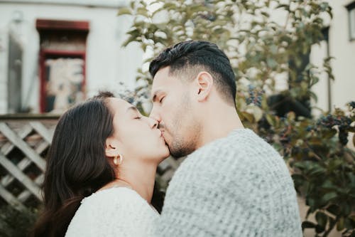 Free Side view of young couple in casual clothes standing in street together in summer day near green plants and building on background and kissing each other tenderly Stock Photo