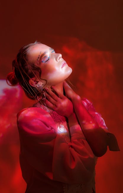 Confident young lady with glimmering eyeshadows in colorful studio covered with red light and shadows with hands on neck and closed eyes