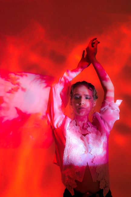 Confident young woman wearing blouse in bright studio covered with red shades and smoke with raised hands and looking away