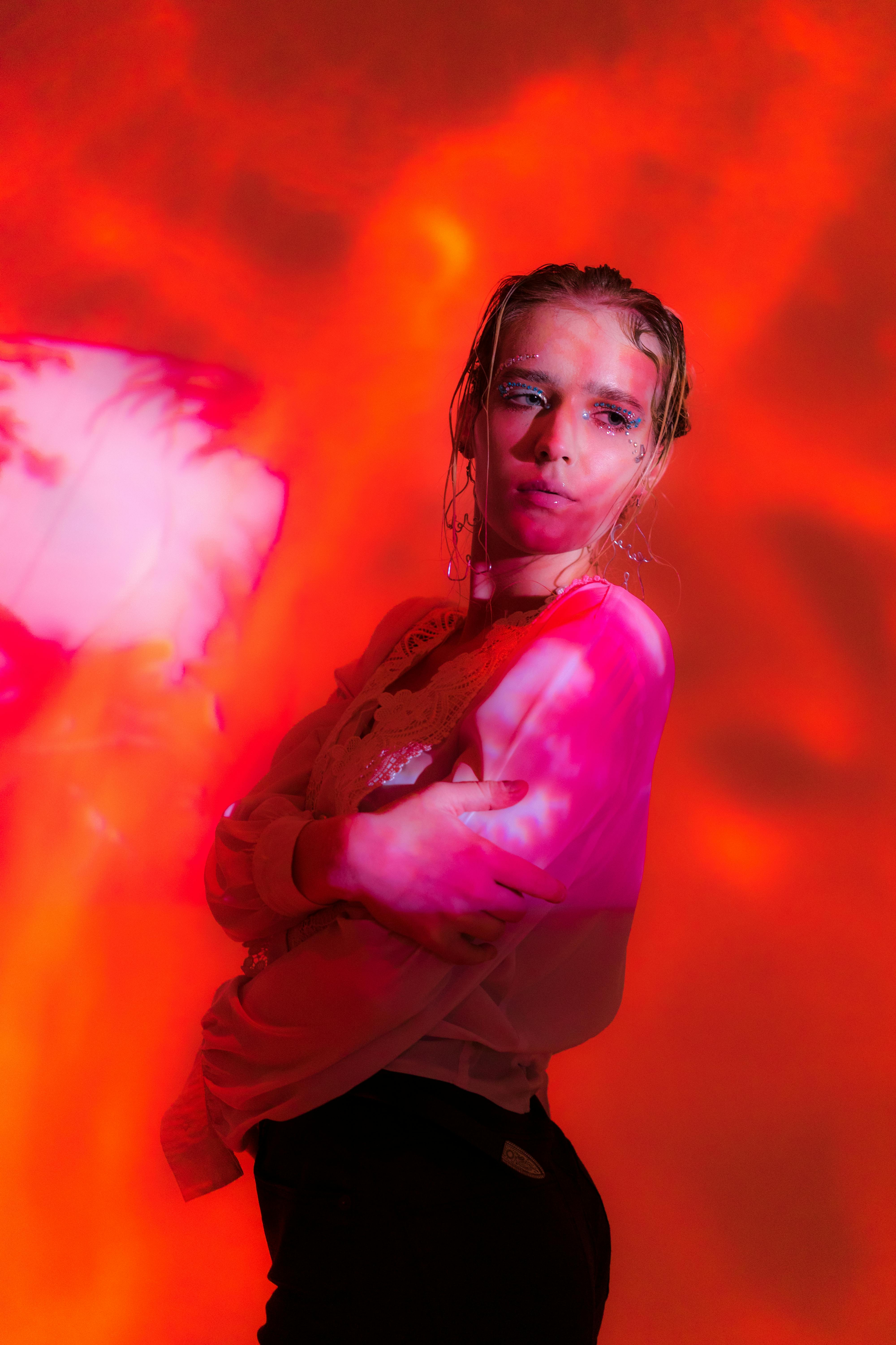 Side view of confident young female in blouse and pants standing in studio covered with red shades and smoke with hand on elbow and looking away