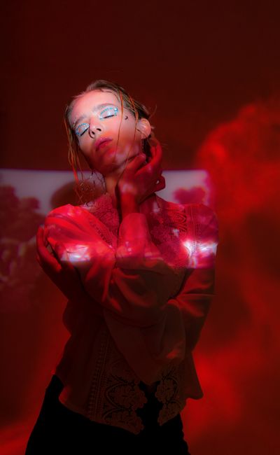Pensive young female with glowing blue eyeshadows in dark studio covered with red shades and lights with hand on neck and closed eyes