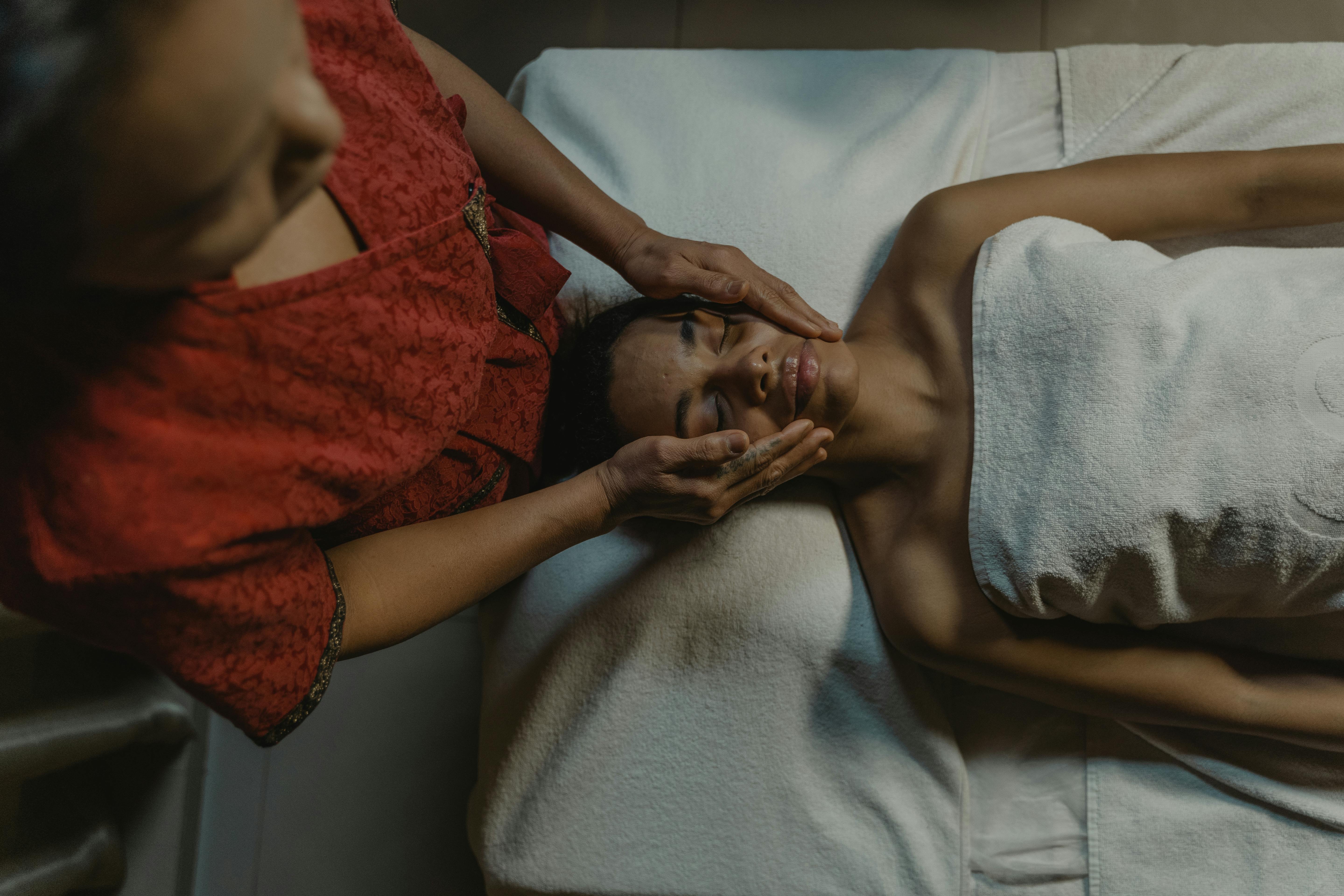 Person receiving a facial massage at a spa while lying on a treatment bed with a towel wrapped around the body