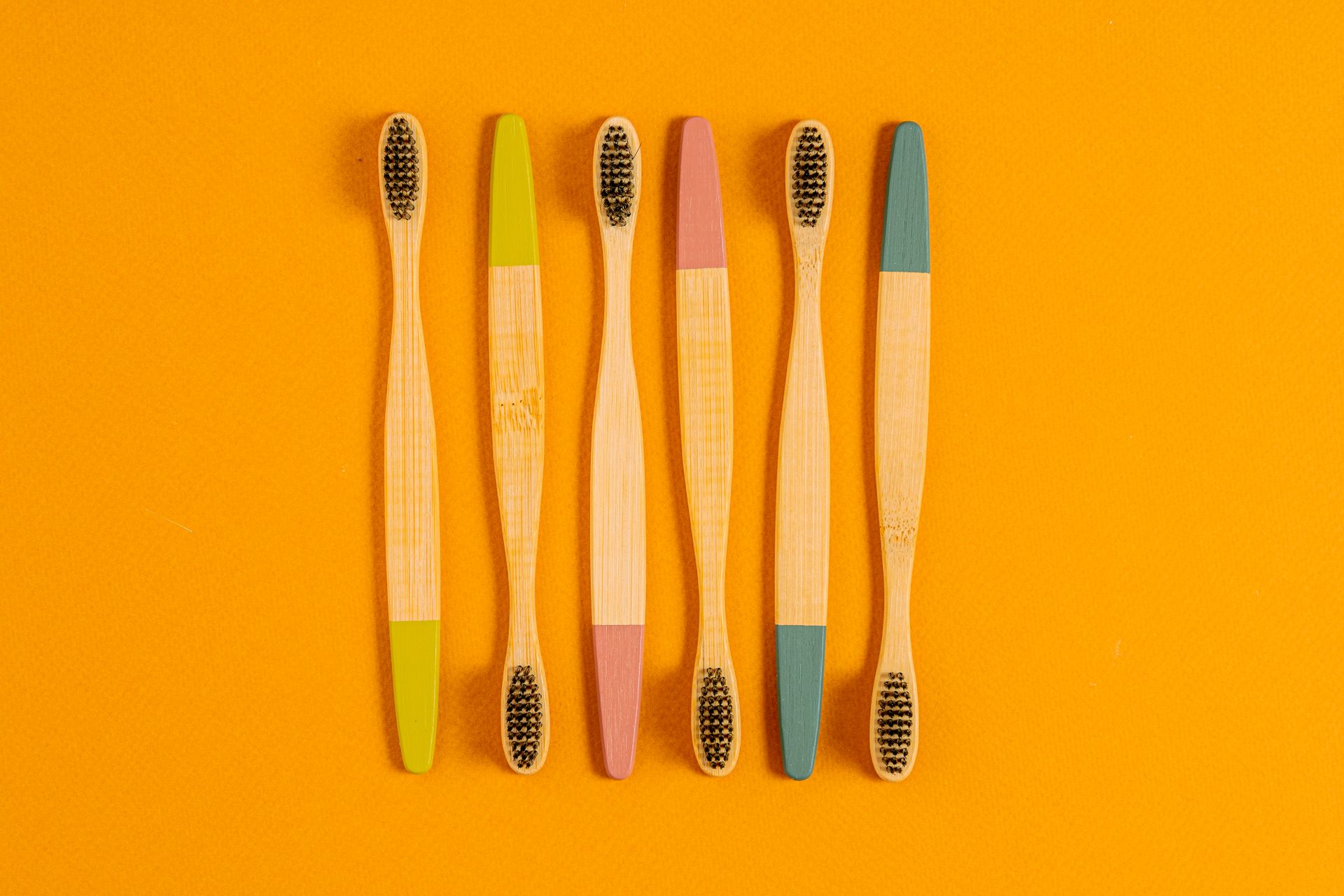 Six bamboo toothbrushes with colorful tips on a vibrant orange surface.