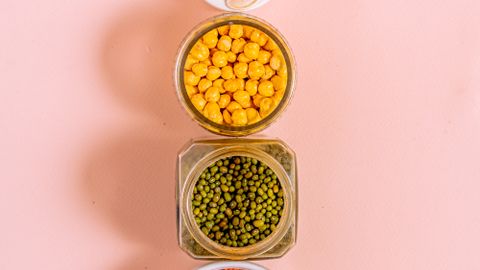 Vertical arrangement of various seeds and grains in bowls on a pink background.