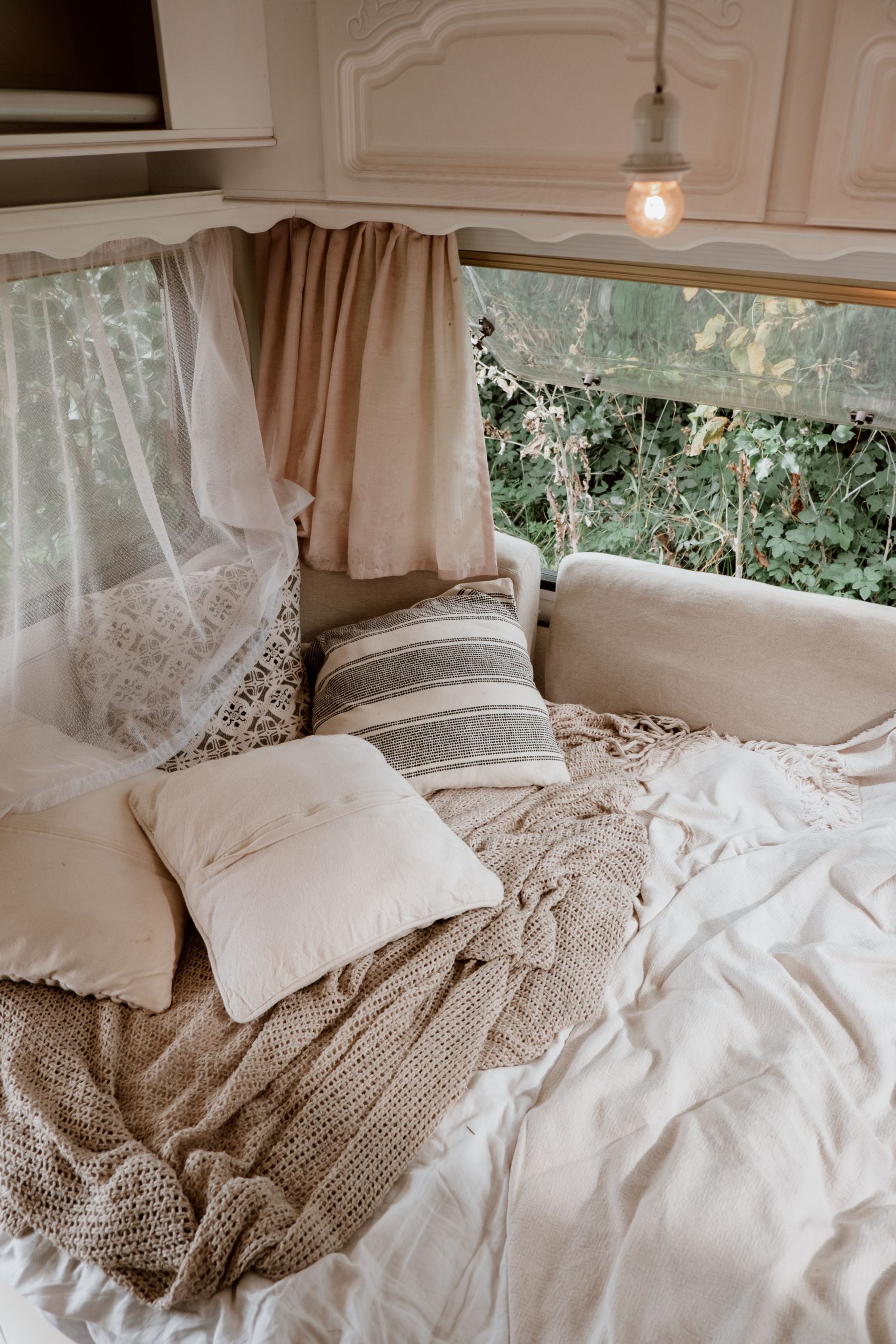 Bed layered with pillows and blankets beside large windows and indoor greenery