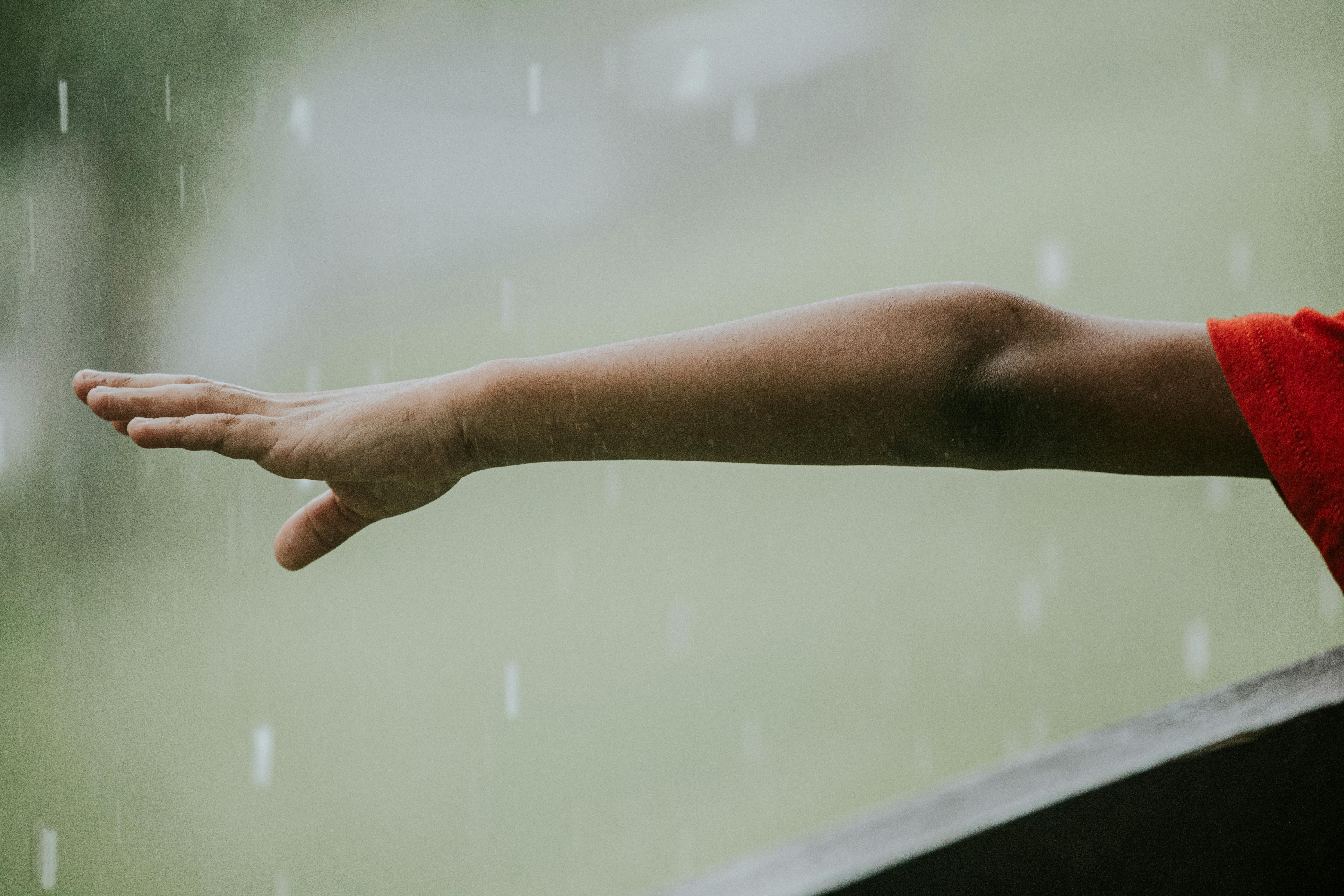 Side view of crop faceless kid in red t shirt near fence pulling hand to rain drops in street in daytime