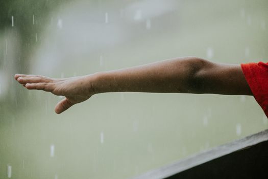 Side view of crop faceless kid in red t shirt near fence pulling hand to rain drops in street in daytime