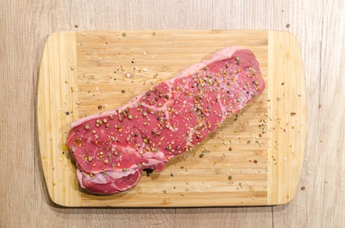 Photographie à Plat D'une Tranche De Viande Sur Le Dessus D'une Planche à Découper Saupoudrée De Grains De Poivre Moulu