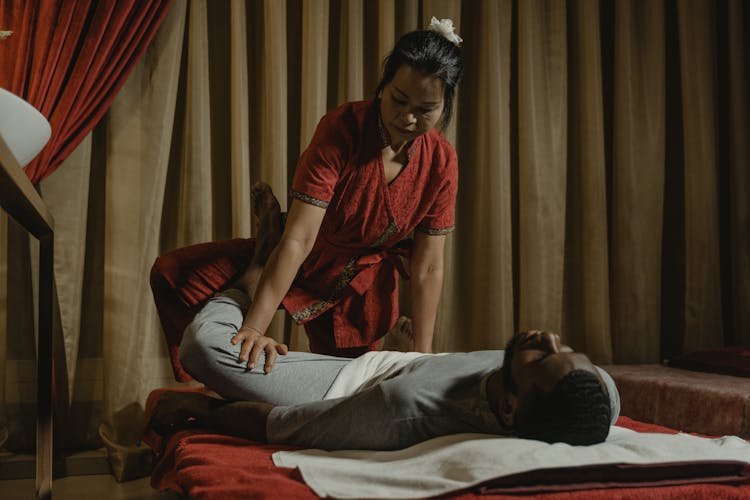 A Woman In Red Shirt Stretching Lying Man's Leg In Massage Room