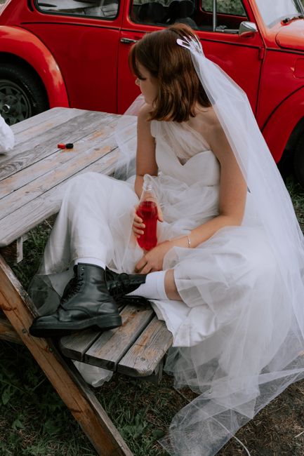 Bride in a wedding gown and black boots sitting on a wooden bench next to a vintage red car, holding a drink.