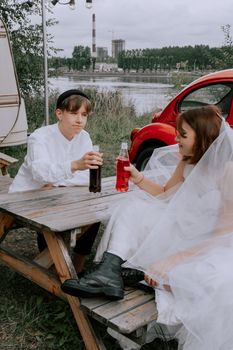 A couple enjoying a casual moment in wedding attire with soda outdoors by a river.
