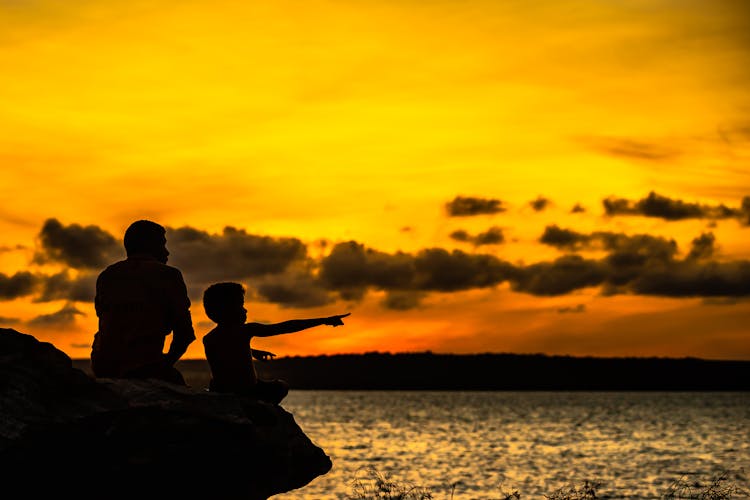 Silhouette Of Man And Boy Pointing Finger While Sitting On Rock Near Body Of Water During Sunset