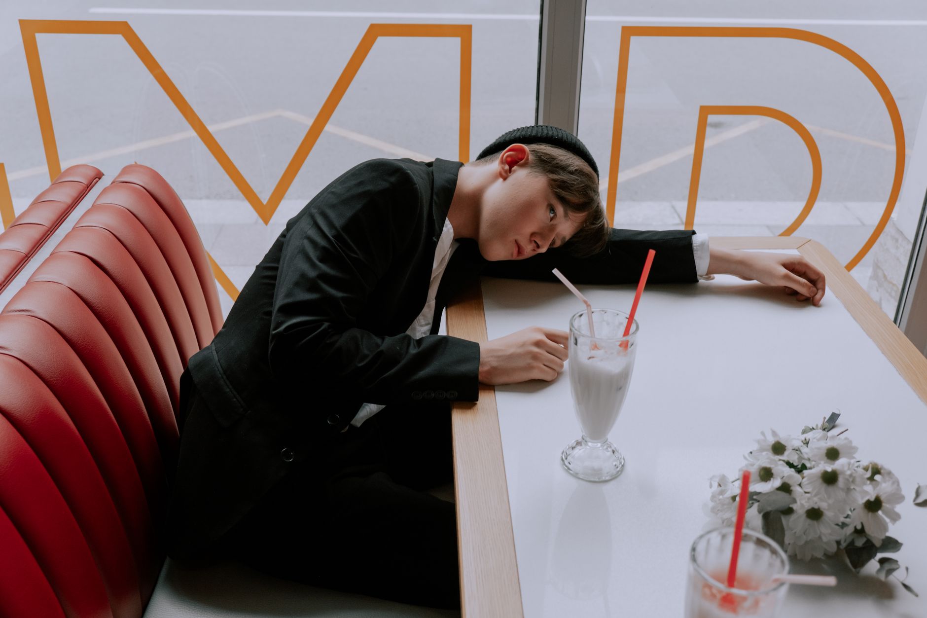 Young man in stylish outfit relaxing at a cafe booth with milkshake and white flowers.