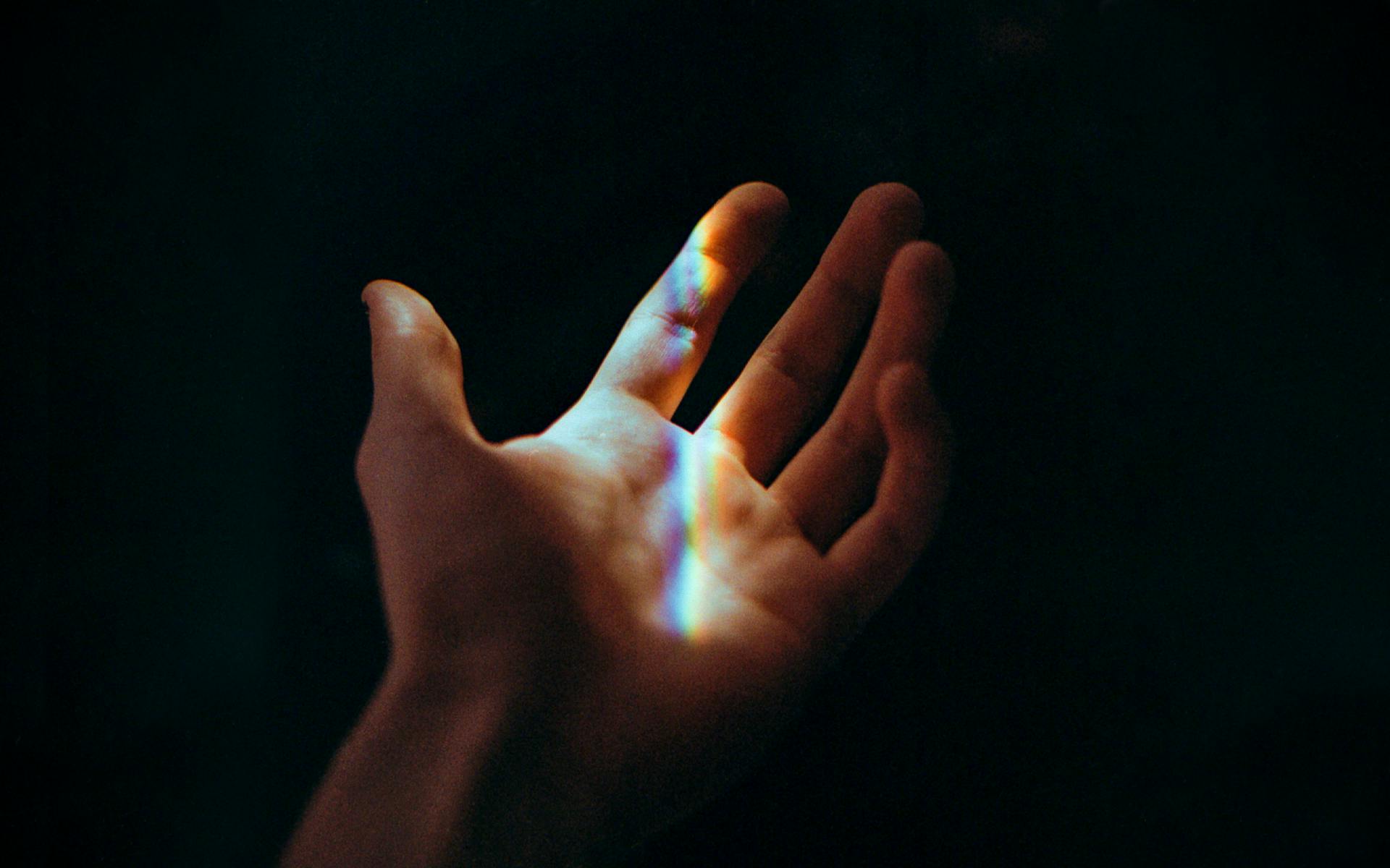 A hand reaching against a dark background with rainbow light, symbolizing hope and mystery.