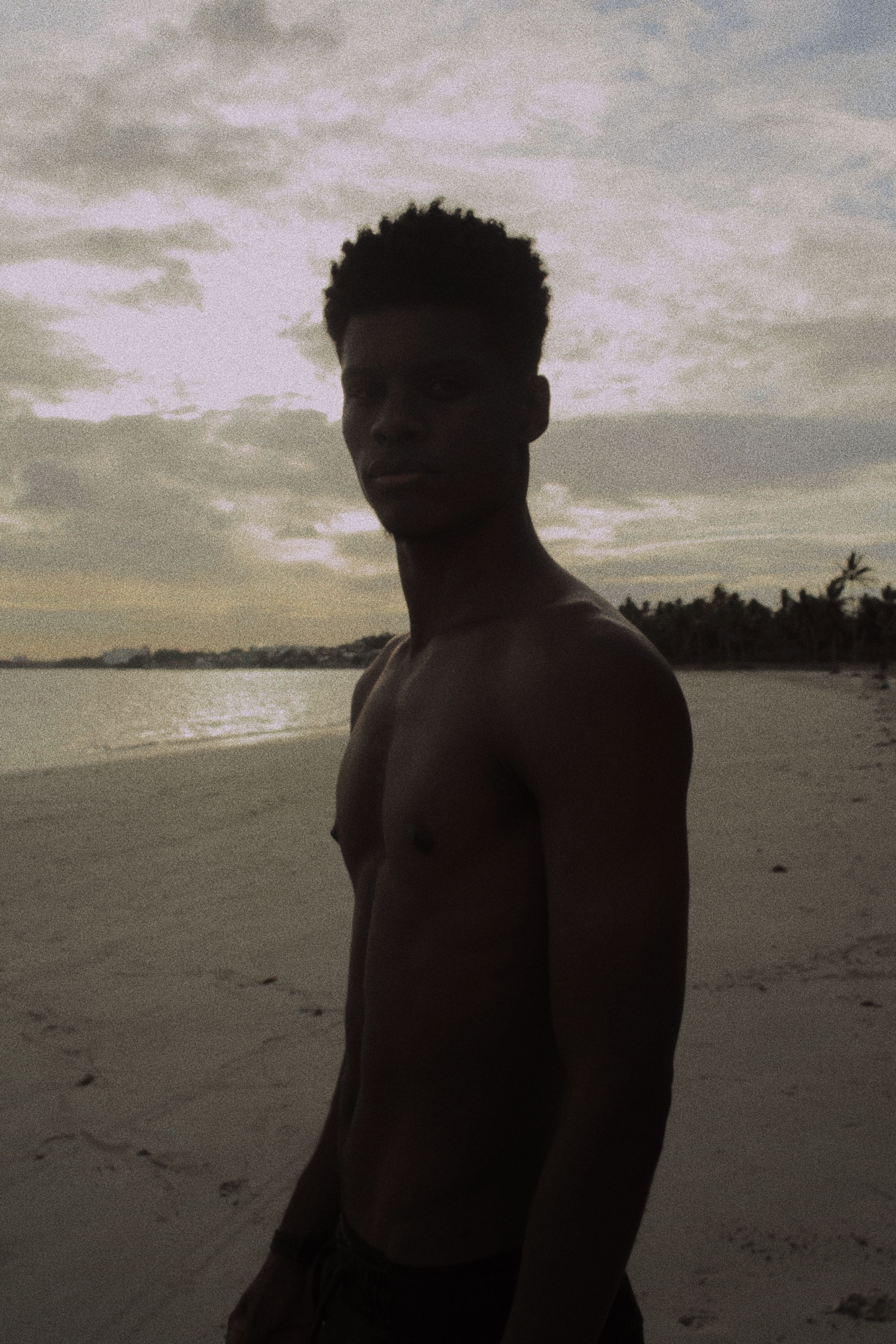 Shirtless man stands on a serene beach at sunset in Madre de Deus, Brazil.