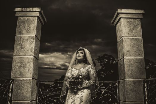 Black and white of dreamy female bride in wedding dress and veil with bouquet of flowers standing near fence and columns near trees under cloudy sky
