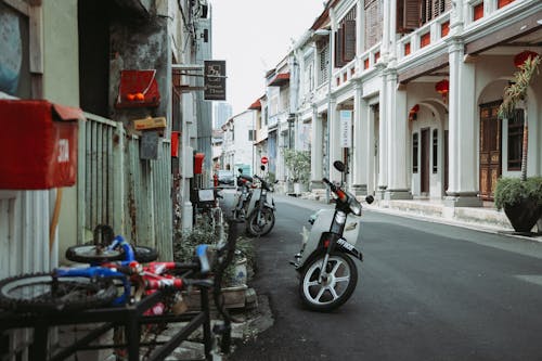 White motorcycles parked on asphalt sidewalk between typical residential buildings near rusty metal fence and bicycle on street in city