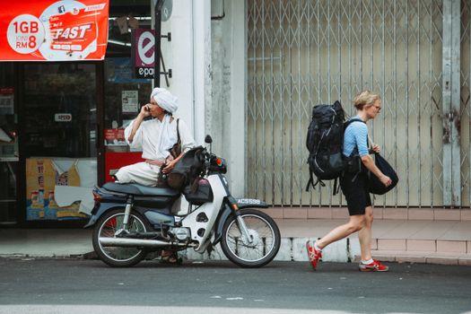 Full body of tourist with backpack walking on sidewalk along building near Indian male in turban on motorcycle on street in city