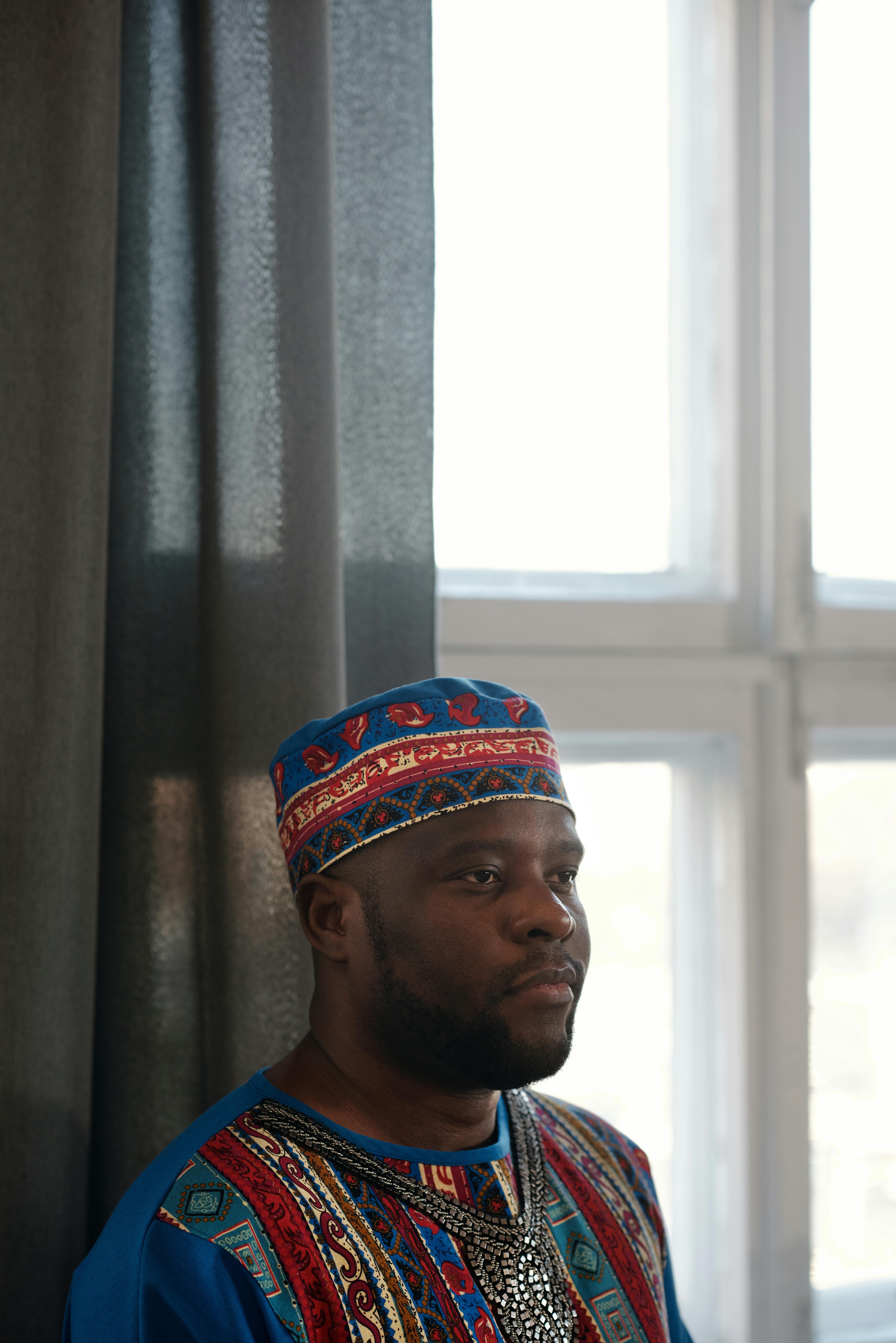An adult man wearing a colorful dashiki and kufi, posing indoors with natural light.