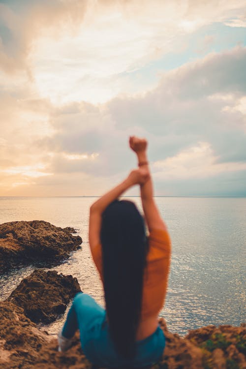 Back view of anonymous female raising arms while resting on rocky coast against endless sea under cloudy sky