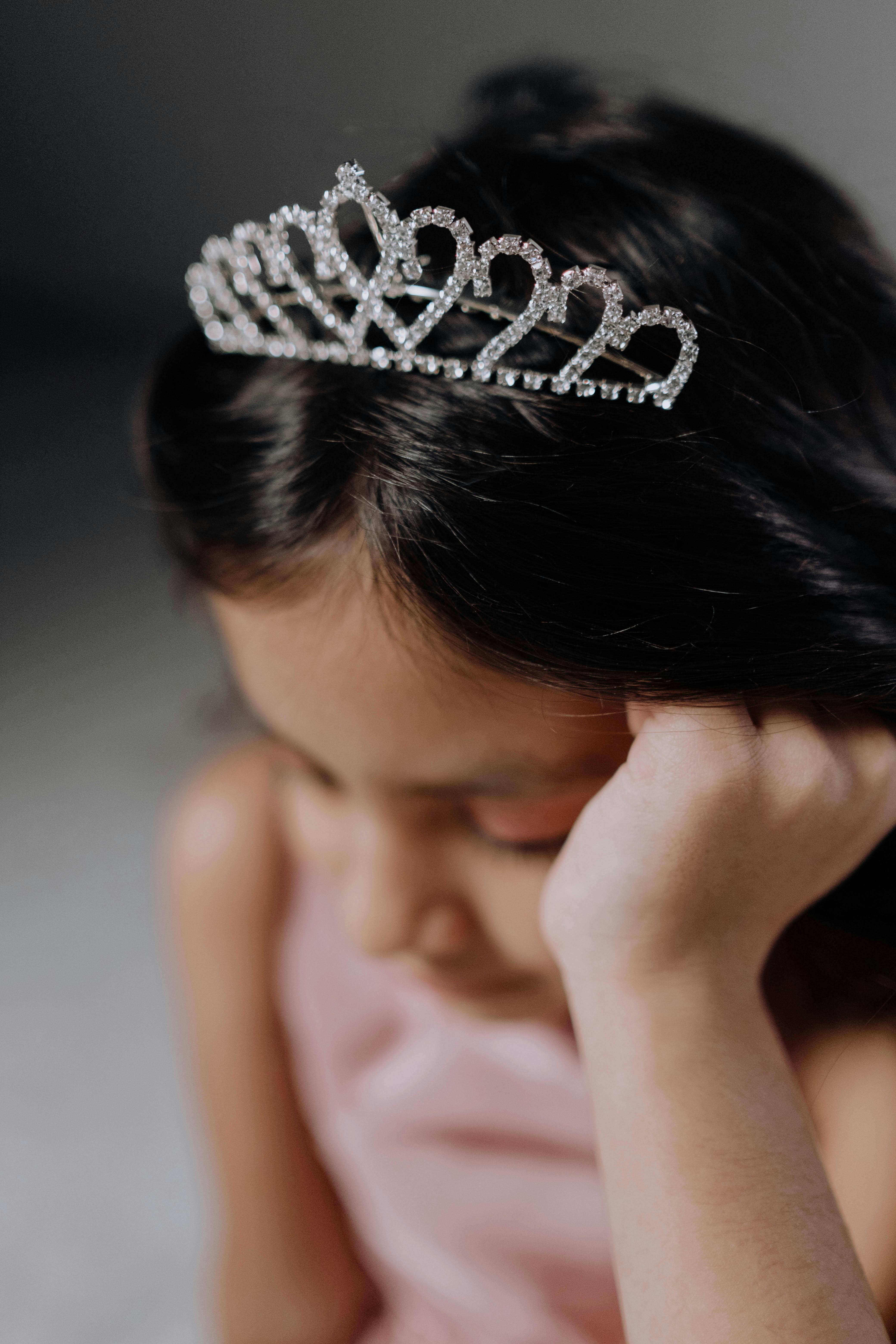 A young girl wearing a tiara, deep in thought indoors.