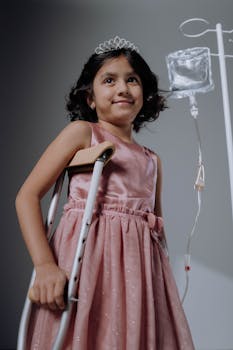 A smiling young girl with a crutch and IV stand in a studio, wearing a pink dress and tiara.
