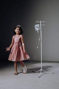 Smiling girl in a pink dress with IV attached, standing in a studio setting.