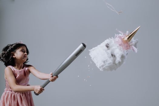 Young girl in a pink dress hits a unicorn piñata indoors with a bat.