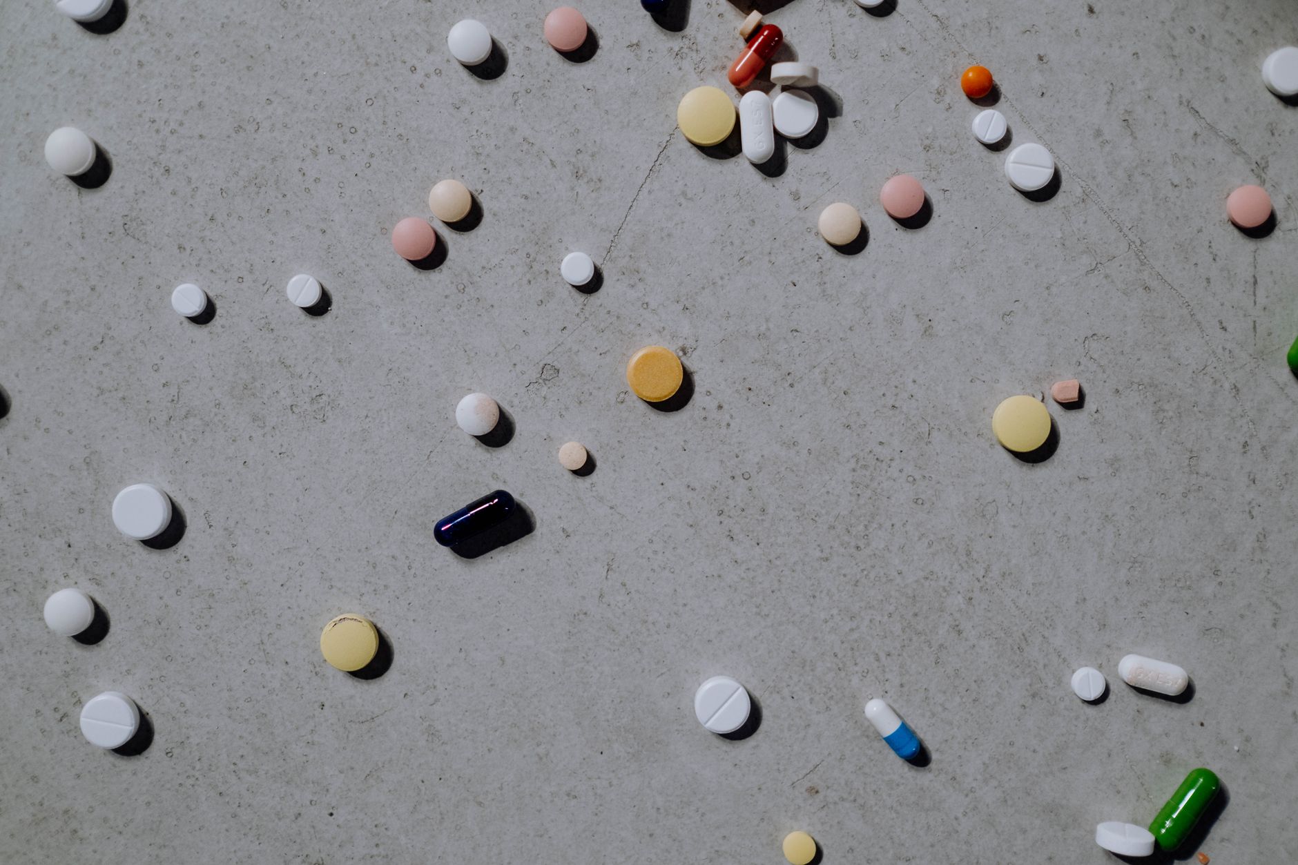 Varied colorful pills and tablets scattered on a grey surface, viewed from above.