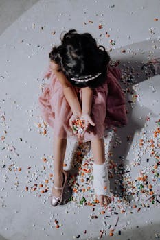 A young girl in a pink dress sits with an orthopedic cast surrounded by various pills and capsules.