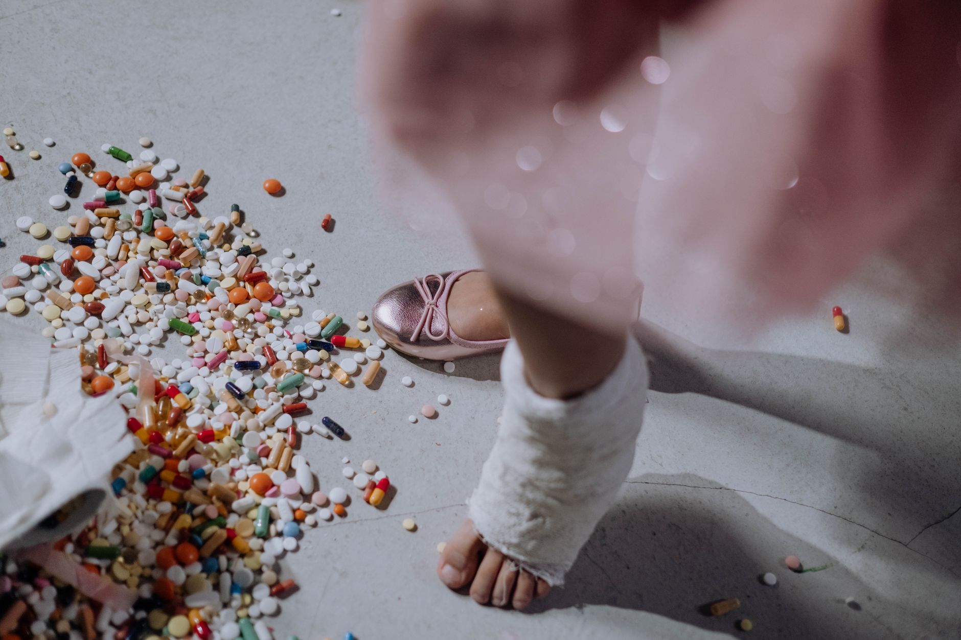 A child's foot in a cast next to a pile of scattered pills on a white floor.