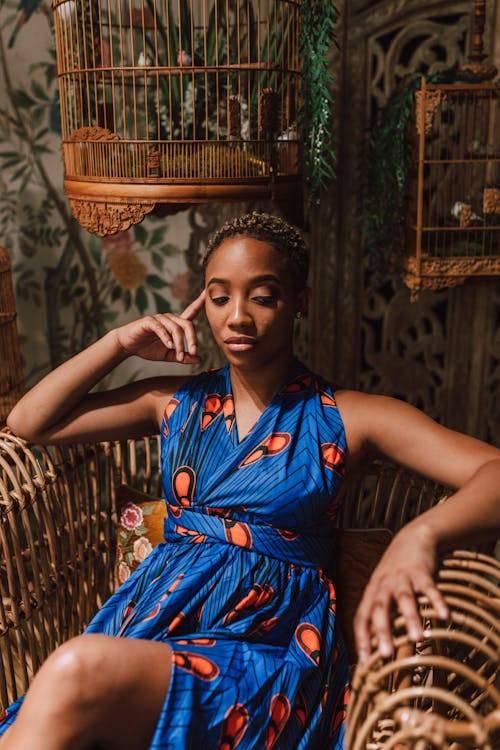 Free Woman in Blue and Yellow Floral Sleeveless Dress Sitting on Brown Woven Chair Stock Photo