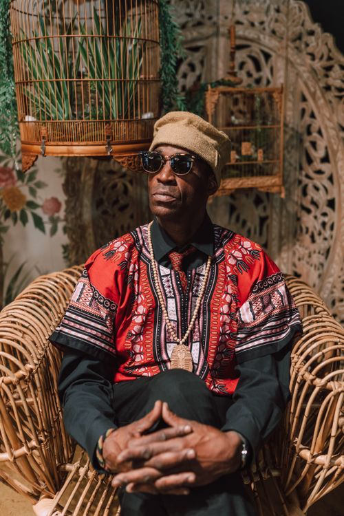 Free Portrait of an African man in vibrant dashiki and kufi, seated with birdcages in the background indoors. Stock Photo