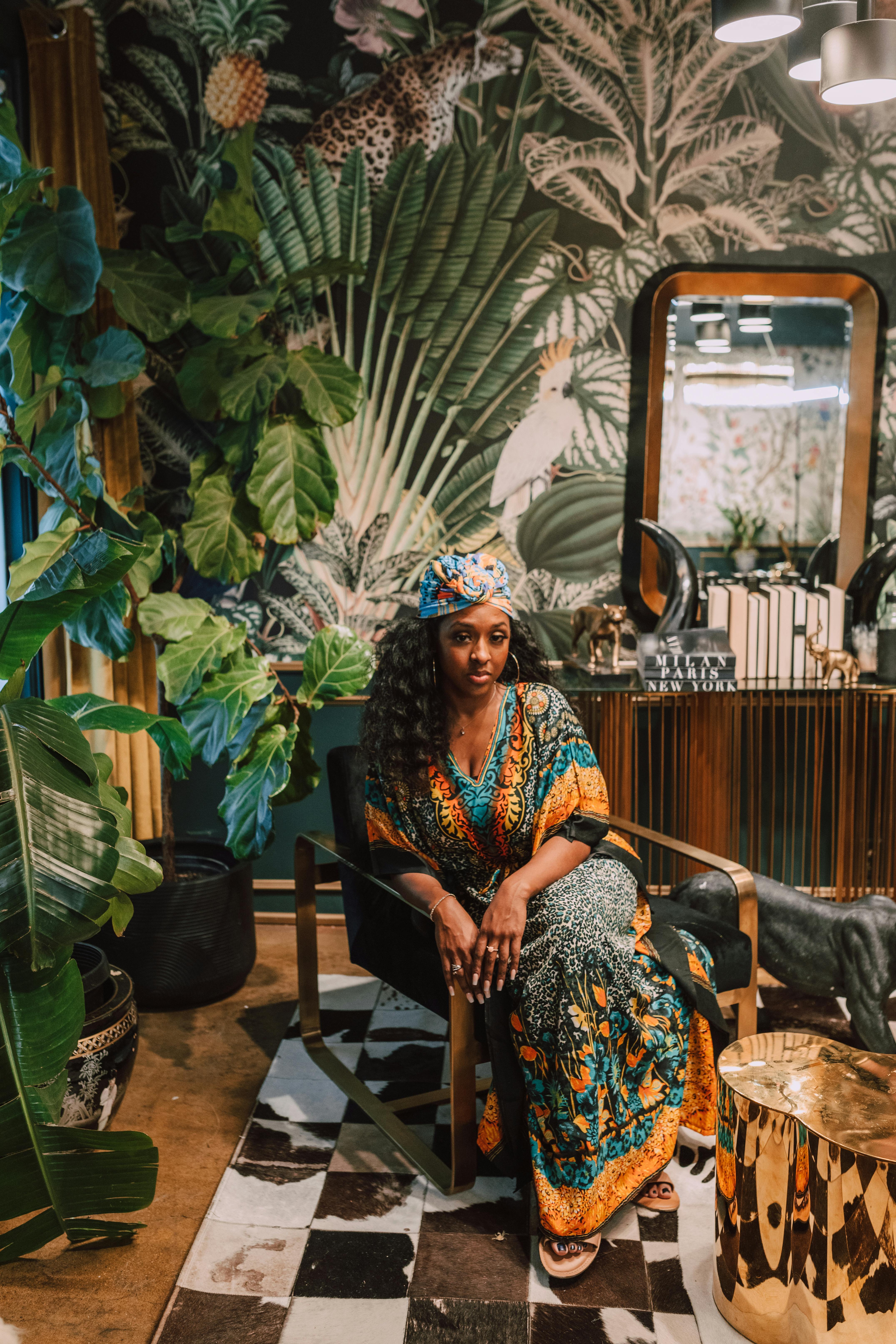A stylish woman poses gracefully in a colorful kaftan, surrounded by lush indoor foliage and elegant decor.
