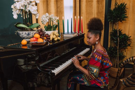 Woman in traditional attire celebrating Kwanzaa playing the piano indoors.