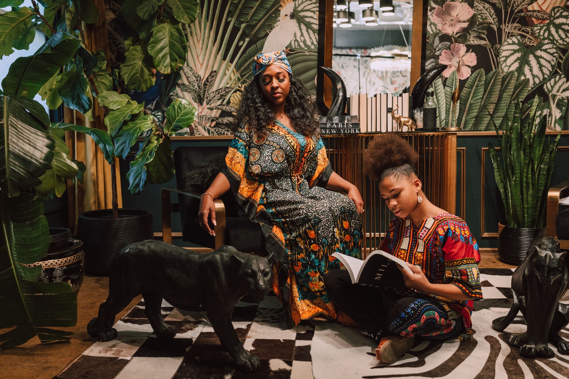 Two women in vibrant African print dresses relaxing among lush indoor plants and decor.