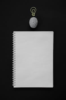 Vertical flat lay of a blank notebook and a brain with a light bulb symbol on black surface, ideal for creativity themes.