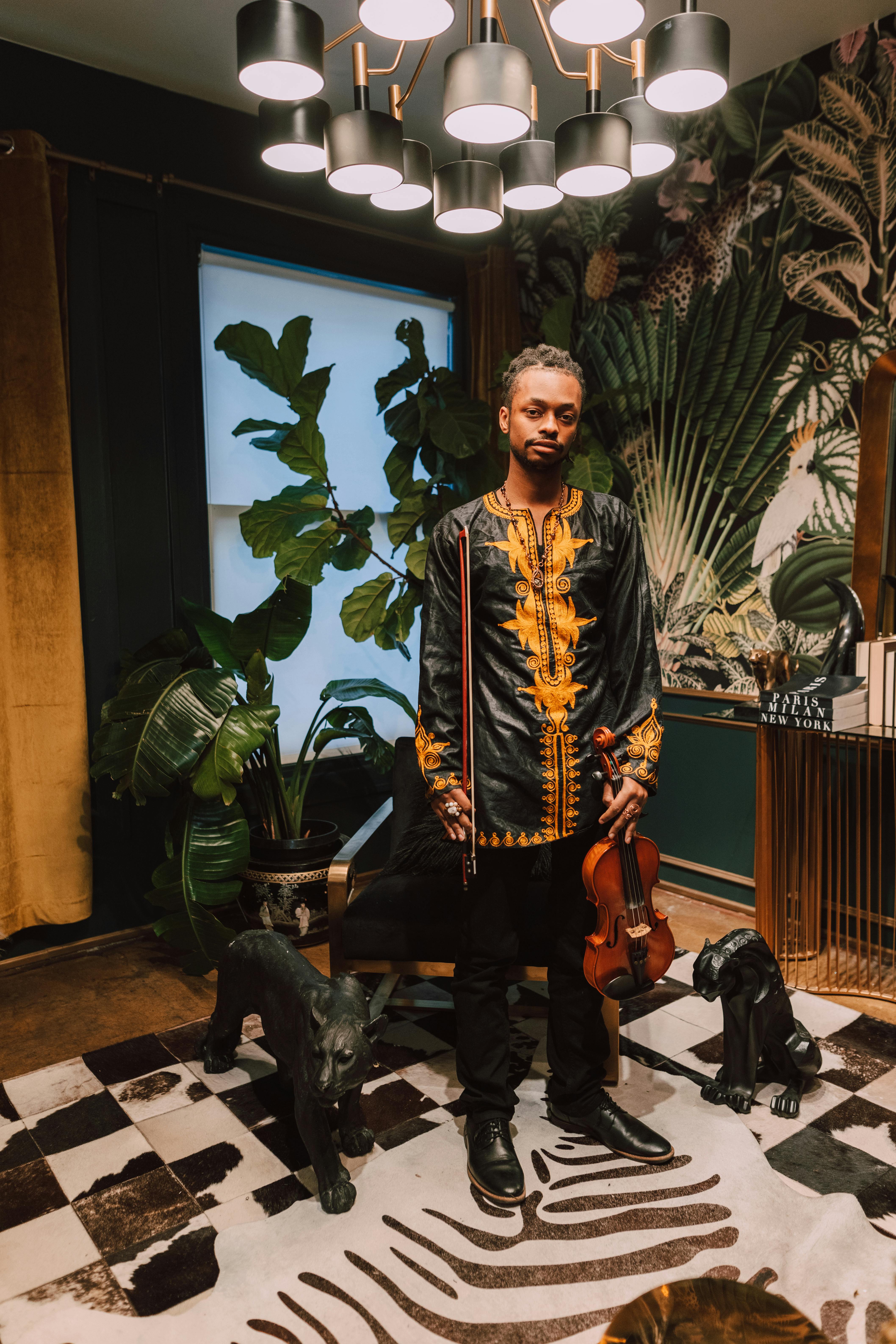 Man in a black-gold dashiki holds a violin in a stylish room with plants and art.