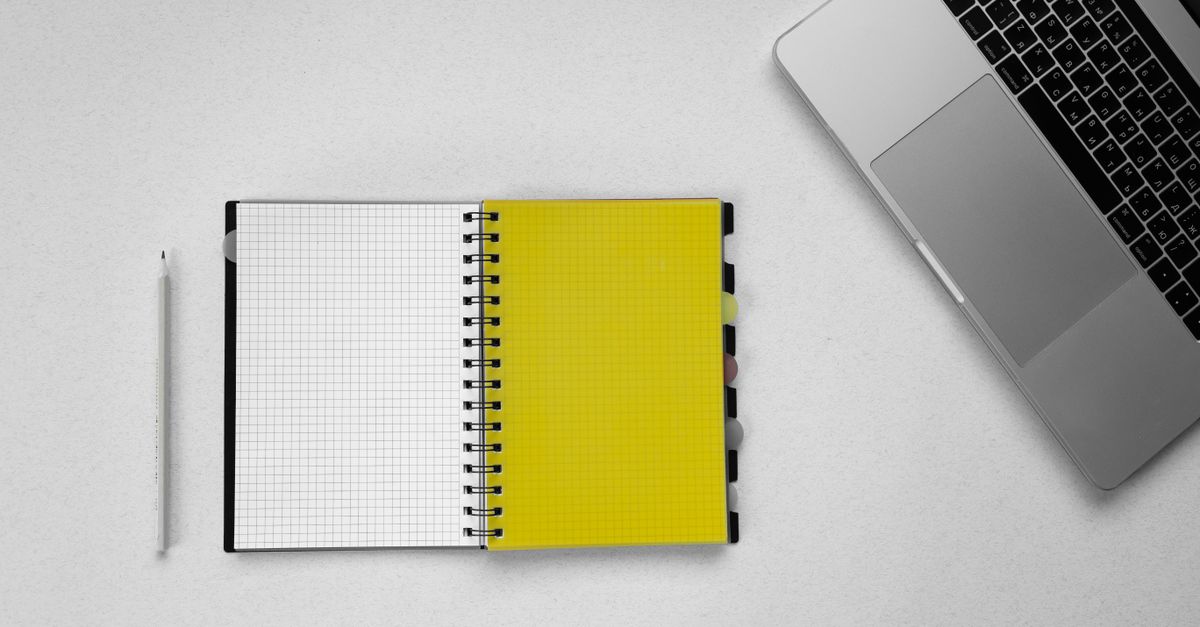 Top view of a modern workspace with a laptop, open notebook, and a pencil on a white surface.