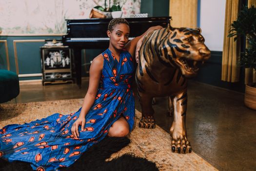 A stylish woman in a vibrant blue dress poses indoors with a decorative tiger statue, exuding elegance.