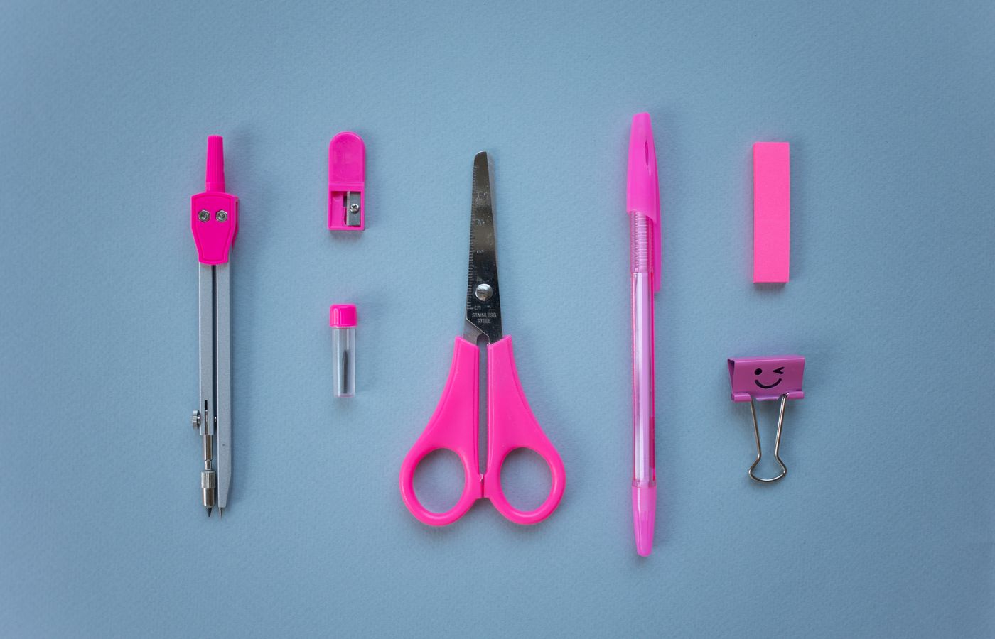 Flat lay of vibrant pink school supplies on a baby blue background, including scissors, pen, and compass.