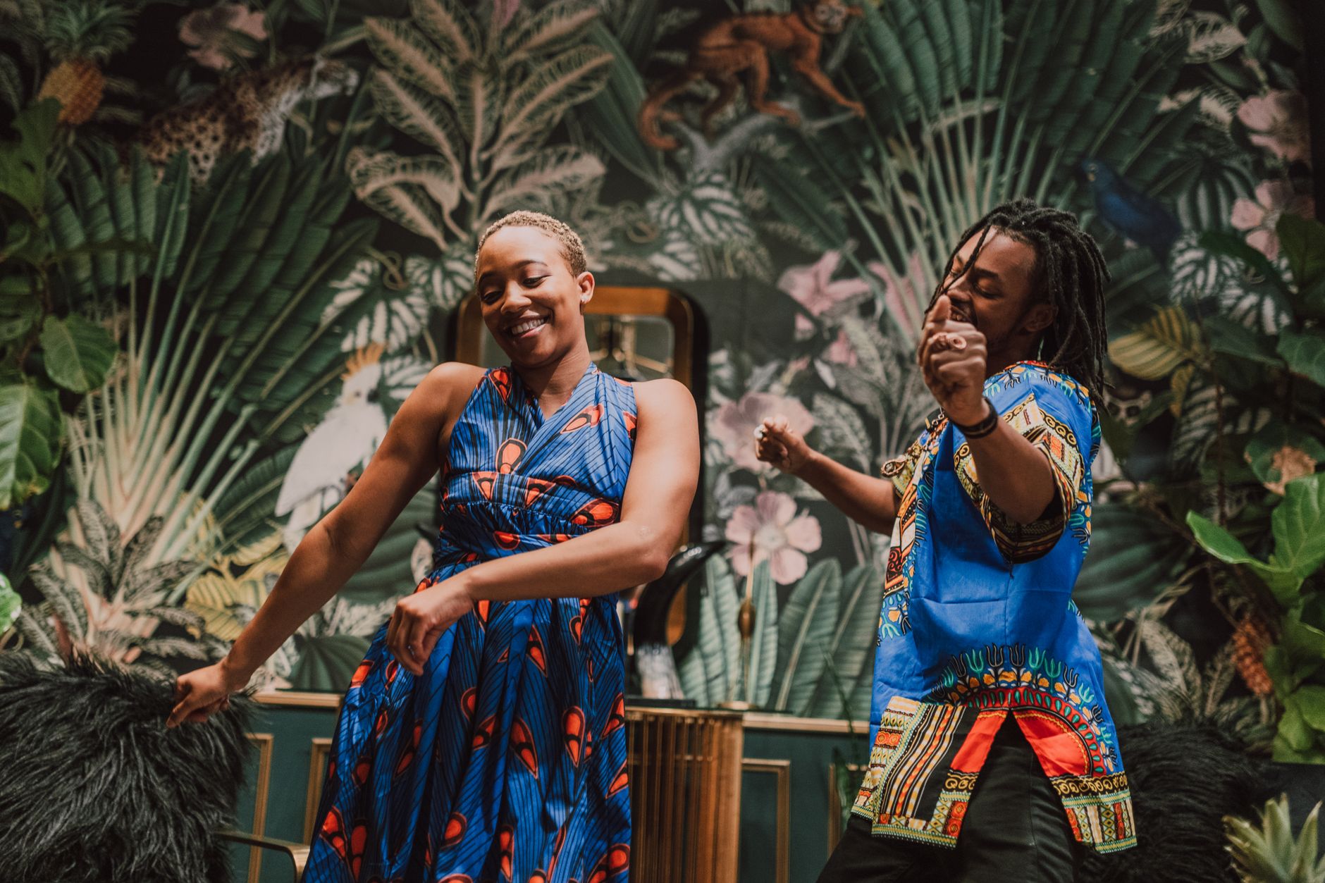 Two adults dancing joyfully in colorful traditional African clothing with a lush backdrop.