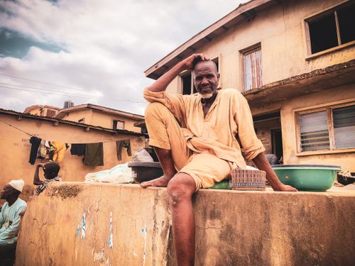 Free Elderly man in casual attire sitting on a wall outdoors with houses in the background. Stock Photo