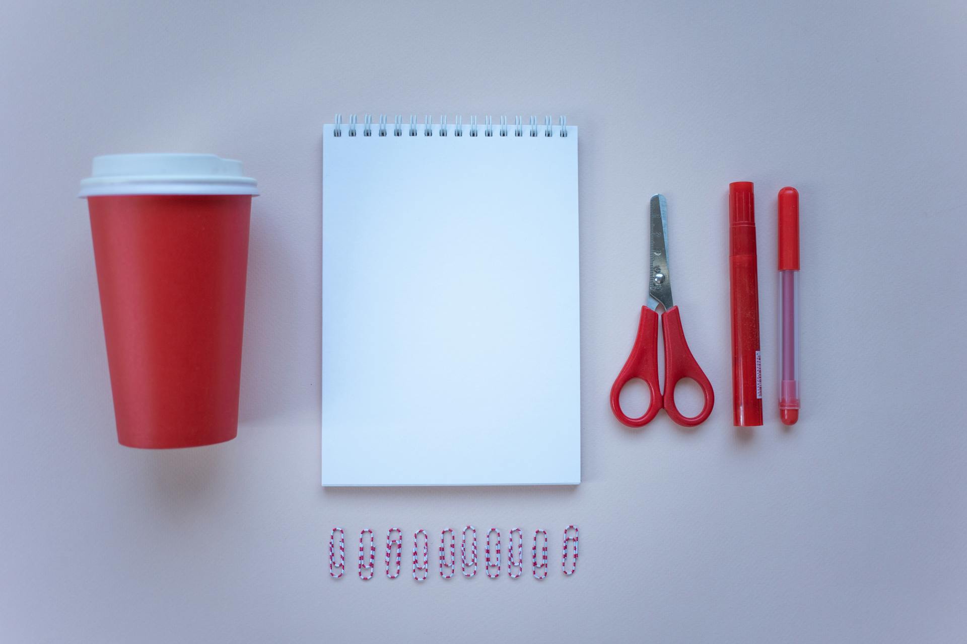 Flat lay of red and white school supplies, including notepad, scissors, and paper cup, on a neutral background.