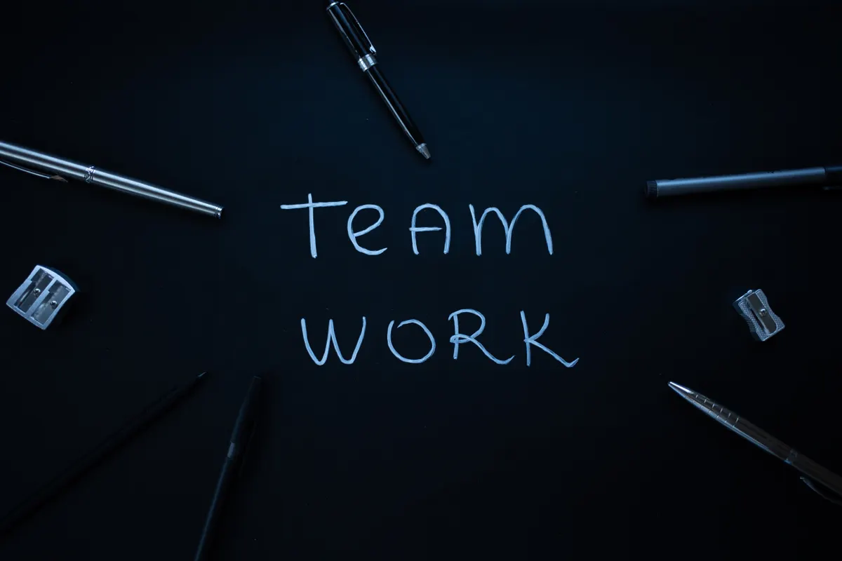 The words 'TEAM WORK' written in white chalk on a black surface, surrounded by pens and sharpeners, symbolizing human-AI collaboration for content creation.