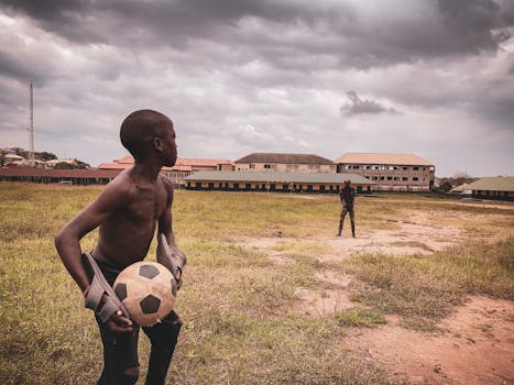 Two boys playing soccer on a grassy field with buildings in the background under cloudy skies.
