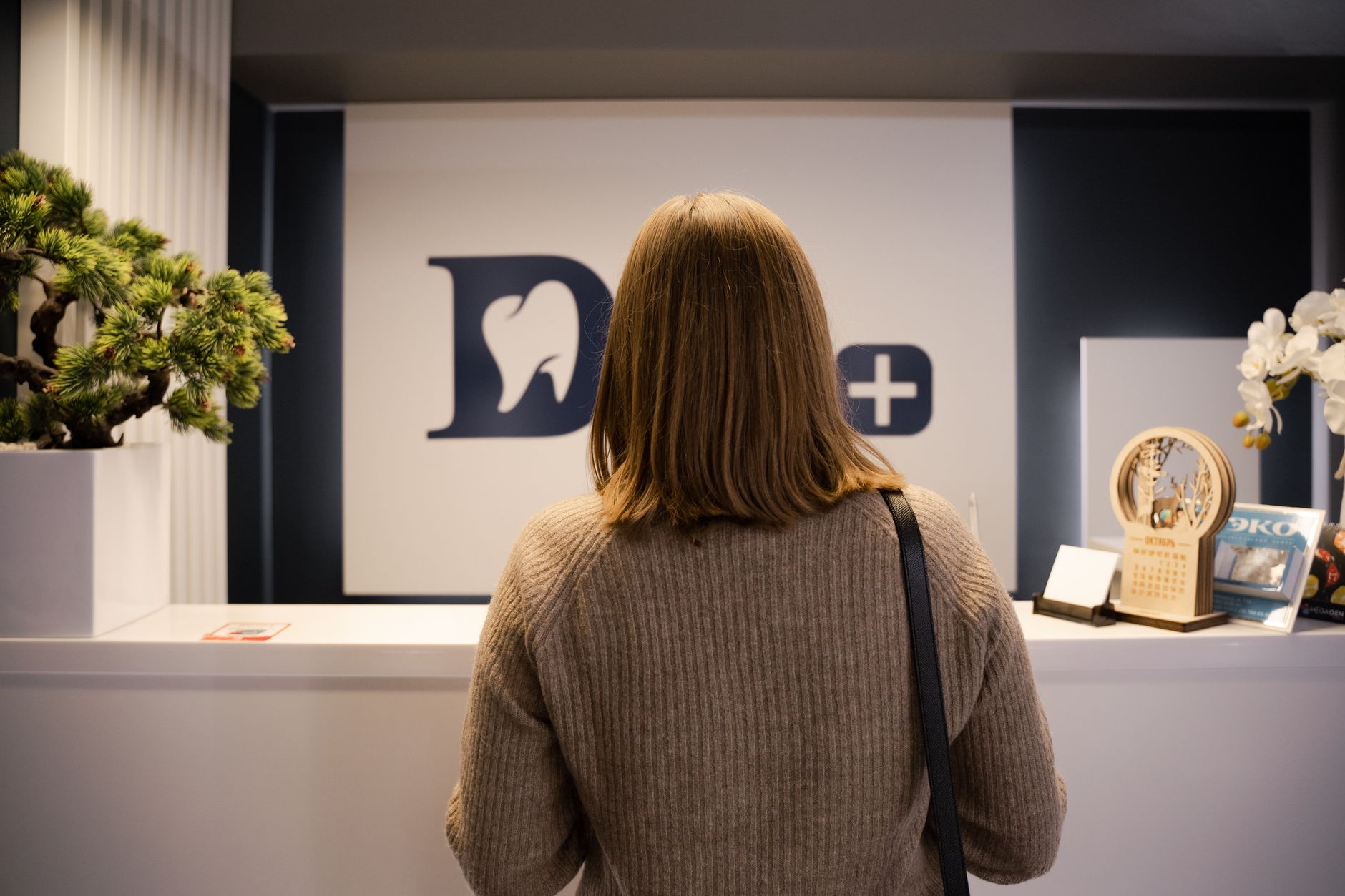 A woman stands at the dental clinic reception wearing a brown sweater, facing away.